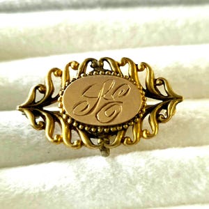 May include: An oval gold brooch featuring an intricate filigree border with scrolling metalwork. The center of the brooch contains a smooth, flat oval plate engraved with the cursive letters "GC" surrounded by a small beaded edge detail.