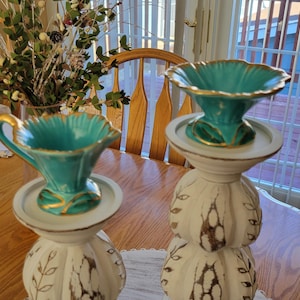 May include: Two decorative teal and gold-trimmed ceramic vases with a scalloped rim, set atop ornate white pedestals. The vases have a unique shape and are accented with gold detailing. The pedestals have a textured, floral design.