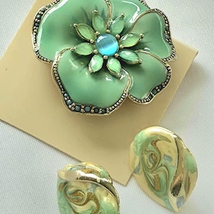 May include: A large, green flower brooch with a blue center stone and gold-colored trim. Two matching earrings with a swirl design in green, yellow, and gold are also present. The jewelry is set on a neutral background.