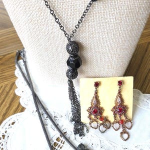 May include: A long necklace featuring a dark metal chain with black beads and a tassel pendant, paired with a separate mesh chain necklace with dark gray crystals and a pair of ornate drop earrings with red stones on a beige display card.