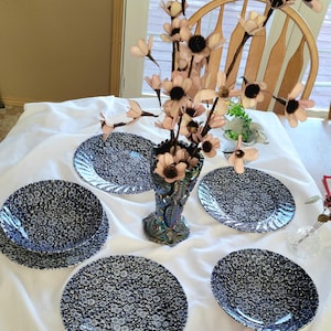 May include: A table setting featuring a collection of dark blue and white floral patterned plates and bowls. A decorative iridescent vase holds a bouquet of pink flowers. The setting is on a white tablecloth.