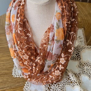 May include: An infinity scarf featuring a sheer brown mesh fabric layered with a white floral print. The scarf is embellished with embroidered orange and brown flowers, delicate white vine patterns, and small clear sequins throughout the design.