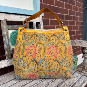 May include: A yellow tote bag featuring a floral design in pink, blue, and green. It has a brown handle and yellow leather details. The bag is placed on a weathered wooden bench.