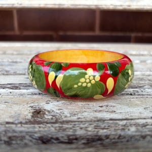 May include: A round bangle bracelet featuring a vibrant red base colour with hand-painted green leaf motifs and small yellow dot clusters. The interior of the circular accessory is a solid, smooth yellow finish, contrasting with the exterior floral pattern.