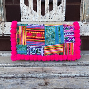 May include: A rectangular clutch bag featuring a patchwork of vibrant, multi-coloured woven fabric panels with intricate geometric patterns. The edges of the bag are decorated with a thick border of bright hot pink pom-poms on three sides.