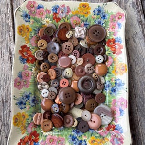 May include: A selection of mixed-size buttons in brown, beige, and cream tones, arranged on a floral patterned plate. The plate has a cream base with vibrant flower designs.