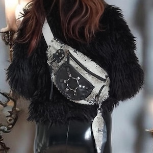 Pode incluir: Um casaco de pelo sintético preto, leggings pretas brilhantes e botas pretas com recortes, combinados com uma pochete com padrão de pele de cobra em branco e preto, com um emblema circular de couro preto com tachas prateadas e um pendente.