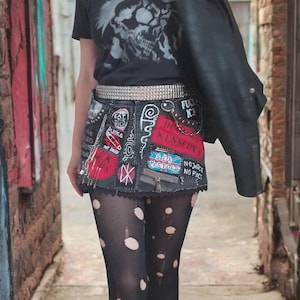 Può includere: Una maglietta nera con grafica a teschio, abbinata a una minigonna in denim decorata con toppe e una cintura borchiata. L'outfit include collant strappati e stivali neri. Una giacca di pelle nera è tenuta sulla spalla. Il testo "Sweet Iron Skirt" è in basso.