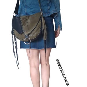 May include: A woman wearing a denim jacket and a short denim skirt is carrying a green and black leather crossbody bag with fringe details. The bag has a silver ring on the front.