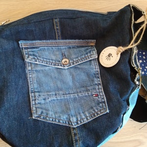 Puede incluir: Un bolso tote de mezclilla azul con un bolsillo grande en la parte delantera. El bolso est&aacute; hecho de materiales reciclados y tiene una etiqueta de cuero con el texto "Universo Etico" y una huella de mano.