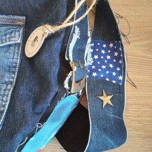 Puede incluir: Un bolso de mezclilla azul con un parche de tela azul y blanco con estampado de estrellas, una estrella dorada y un asa de cuerda marr&oacute;n. El bolso tiene una hebilla de metal y un detalle de tela a rayas azul y blanco. El bolso est&aacute; etiquetado como "justine.art".