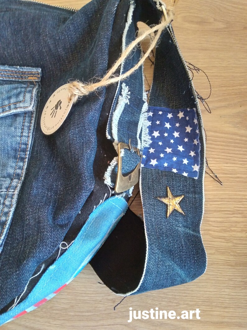 Puede incluir: Un bolso de mezclilla azul con un parche de tela azul y blanco con estampado de estrellas, una estrella dorada y un asa de cuerda marr&oacute;n. El bolso tiene una hebilla de metal y un detalle de tela a rayas azul y blanco. El bolso est&aacute; etiquetado como "justine.art".