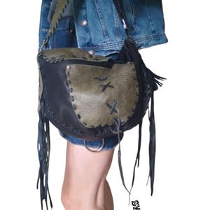 May include: A black and green leather shoulder bag with fringe details. The bag has a flap closure and a strap that can be adjusted. The bag is worn by a person wearing a denim jacket.