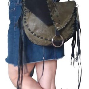 May include: A green, black, and brown leather crossbody bag with fringe detailing. The bag has a silver ring closure and a black strap. The bag is worn by a person wearing a denim skirt. The text "SWEET IRON BAGS" is visible in the bottom right corner of the image.