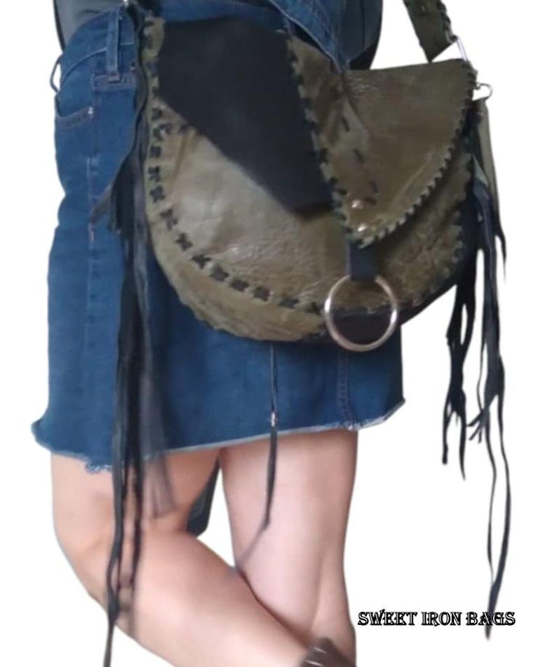 May include: A green, black, and brown leather crossbody bag with fringe detailing. The bag has a silver ring closure and a black strap. The bag is worn by a person wearing a denim skirt. The text "SWEET IRON BAGS" is visible in the bottom right corner of the image.
