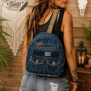 May include: A dark blue denim backpack featuring a leopard print strap, two front pockets with metal stud accents, and a brown leather-style brand patch labeled Sweet Iron Bags. The bag has a zipper closure and visible stitching throughout.