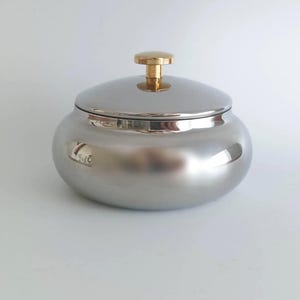 Può includere: Una ciotola in metallo color argento lucido con un corpo arrotondato a profilo basso e un coperchio abbinato. Il coperchio presenta una piccola maniglia a pomello cilindrica color oro centrata sulla parte superiore. La superficie è liscia.