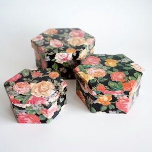 Puede incluir: Tres cajas hexagonales con tapas. Las cajas están decoradas con un estampado floral de rosas rosas, melocotón y amarillas sobre un fondo negro. Las cajas son de diferentes tamaños, la más grande en la parte trasera y la más pequeña en la parte delantera.