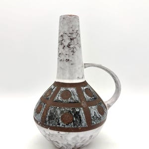May include: A ceramic vase with a bulbous base and a tall, slender neck. The base features a brown and black geometric pattern with circular accents. The handle is curved. The vase has a white, textured finish.
