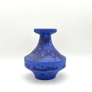 May include: A ceramic vase featuring a vibrant blue glaze with darker blue mottled patches. The vessel has a wide, rounded body that tapers into a narrow neck, ending in a flat, circular rim. The surface texture appears matte and slightly uneven.