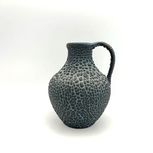 Op de afbeelding: Een blauwe en zwarte keramische vaas met een getextureerd oppervlak en een handvat. De vaas heeft een rond lichaam en een smalle hals.