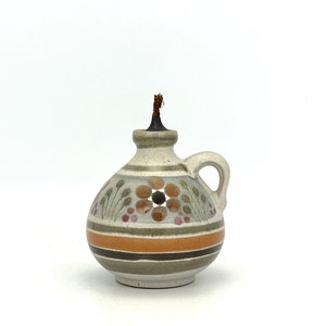 May include: A small, ceramic oil lamp with a brown wick. The lamp is decorated with a floral pattern in shades of brown, green, pink, and white. The lamp has a handle and a round body.