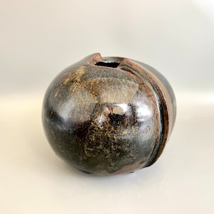 Könnte beinhalten: Eine runde, dunkelbraune Keramikvase mit einem einzigartigen, asymmetrischen Design. Die Vase hat eine glänzende Oberfläche mit Streifen aus hellem Braun und Beige sowie eine rechteckige Öffnung oben. Die Gesamtform ist kugelförmig mit einer vertikalen Nut.