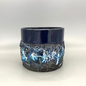 Puede incluir: Un macetero de cerámica azul y blanco con una superficie texturizada y rugosa. El macetero tiene un esmalte azul oscuro en la mitad superior y un esmalte blanco y azul en la mitad inferior.
