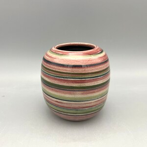 Puede incluir: Un jarrón de cerámica redondo con rayas horizontales en tonos rosa, verde, rojo y azul. El jarrón tiene una abertura ancha y un interior oscuro. Las rayas están espaciadas uniformemente y envuelven todo el cuerpo del jarrón.