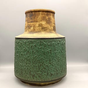 Op de afbeelding: Een keramische vaas met een getextureerd groen onderste gedeelte en een bruin bovenste gedeelte. De vaas heeft een brede basis en een uitlopende bovenkant, met een donkere band die de twee delen scheidt. Het ontwerp is in mid-century modern stijl.