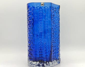 May include: A vibrant blue glass vase with a textured, vertical pattern. The vase has a rectangular shape with rounded edges and a clear base. A small, circular emblem is visible near the top.