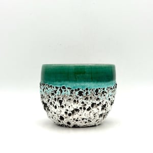 Puede incluir: Una maceta de cerámica con un exterior texturizado y poroso. La parte superior es de un verde intenso brillante, que transiciona a una banda turquesa, y luego a una base moteada blanca y negra. La maceta tiene forma redondeada y está sobre un fondo blanco.