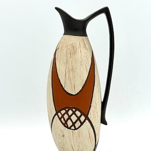 May include: A cream-coloured ceramic jug with a dark brown handle and rim. The jug has a textured surface and is decorated with orange and brown geometric shapes. The design is reminiscent of mid-century modern art.