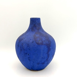 May include: A blue ceramic vase with a textured finish. The vase has a bulbous body and a slender neck. The colour is a deep, saturated blue with darker, mottled patches. The vase is likely handmade and suitable for home decoration.