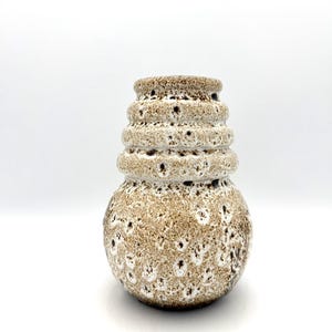 May include: A ceramic vase with a textured, speckled surface in shades of brown and white. The vase has a rounded base and a tiered, cylindrical neck. The design features small, dark spots and a rough, organic aesthetic, suitable for home decor.