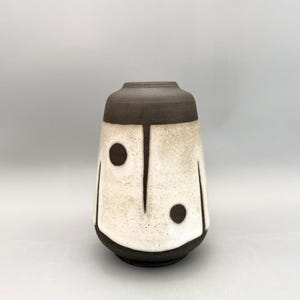 May include: A ceramic vase with a dark brown rim and base, and a cream-coloured body. The body features two dark brown circles and vertical lines, creating a unique face-like design. The vase has a textured surface and a rounded shape.