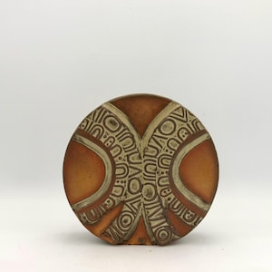 Puede incluir: Un jarrón de cerámica circular con una paleta de colores terracota y crema. La superficie está decorada con intrincados patrones geométricos en relieve y líneas curvas que crean un diseño simétrico en la parte frontal del objeto.