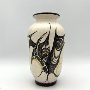 Puede incluir: Jarrón de cerámica blanco con un diseño abstracto en negro y dorado. El jarrón tiene una superficie texturizada y una forma redondeada.