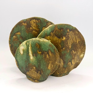 May include: Three circular ceramic sculptures with irregular, wavy edges. The surfaces feature a mottled glaze in shades of forest green, earthy brown, and hints of cream, creating a textured, organic appearance across the three distinct pieces.