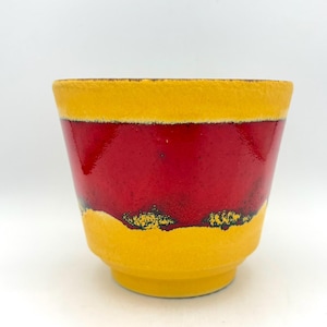 Può includere: Un vaso di ceramica dal design vivace. La parte superiore e inferiore sono gialle, con una fascia rossa lucida al centro. La sezione rossa ha un aspetto strutturato e le sezioni gialle hanno una consistenza leggermente ruvida. Adatto per uso interno o esterno.