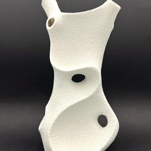 May include: A textured white ceramic vase with an organic, flowing shape. The vase features three circular openings and a narrow spout. The surface has a speckled appearance, and the design is modern and sculptural.