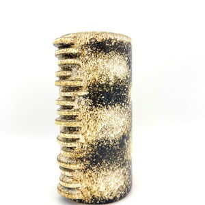 Peut inclure: Un vase cylindrique en céramique présentant un émail moucheté noir et crème. Un côté du récipient affiche une série de crêtes horizontales en relief créant un aspect texturé et nervuré, tandis que le reste de la surface est lisse.