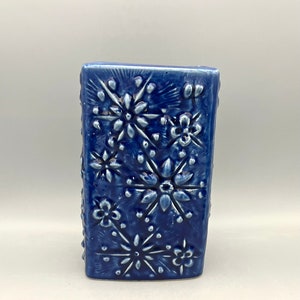 Puede incluir: Un jarrón de cerámica azul con un diseño floral en relieve. El jarrón es de forma rectangular y tiene un acabado liso y brillante.