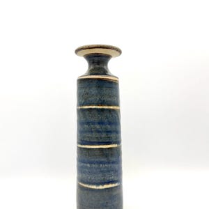 Op de afbeelding: Een hoge, cilindrische keramische vaas met een smalle hals en een uitlopende rand. Het oppervlak is voorzien van een textuurrijk, gevlekt blauw glazuur met horizontale banden van onbedekte lichtbeige klei voor een gelaagde uitstraling.