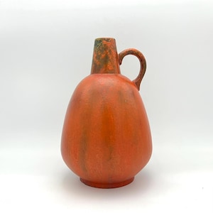 Può includere: Vaso in ceramica arancione con corpo arrotondato e un'unica maniglia. Il vaso ha una smaltatura screziata con sfumature verdi e arancioni.