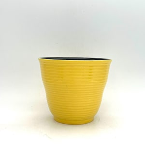 Könnte beinhalten: Ein gelber Keramik-Blumentopf mit geripptem Design. Die Innenseite des Topfes ist schwarz. Der Topf hat eine leuchtend gelbe Farbe und steht auf einer weißen Oberfläche.