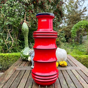May include: A red ceramic vase with black trim and a blue rim. The vase has a cylindrical shape with four raised bands around the body. The vase is sitting on a wooden table.