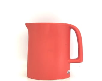 May include: A coral orange ceramic pitcher with a rounded body and a single handle. The pitcher has a small logo on the bottom.