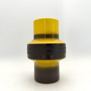 Peut inclure: Un vase en céramique de forme cylindrique, avec un design en blocs de couleurs. Les sections supérieure et inférieure sont jaunes, séparées par une bande texturée brun foncé. Le vase a une finition brillante et est posé sur un fond blanc.
