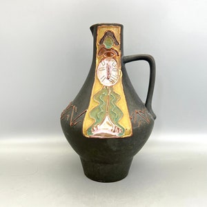 May include: A black ceramic pitcher with a stylized face design in gold, green, and pink. The pitcher has a wide base and a narrow neck with a handle.